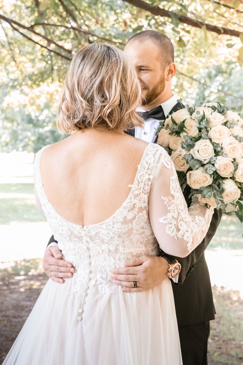 A romantic wedding moment showing a bride in a lace back dress holding a blush rose bouquet embracing her groom outdoors.