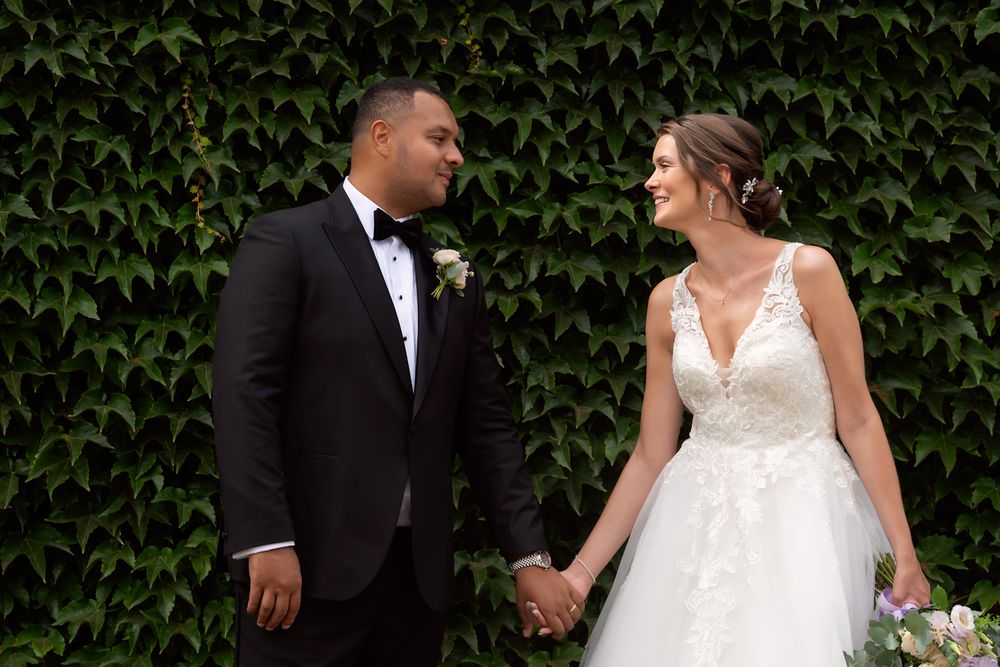 Couple in formal wedding attire walking through lush green ivy covered wall background.