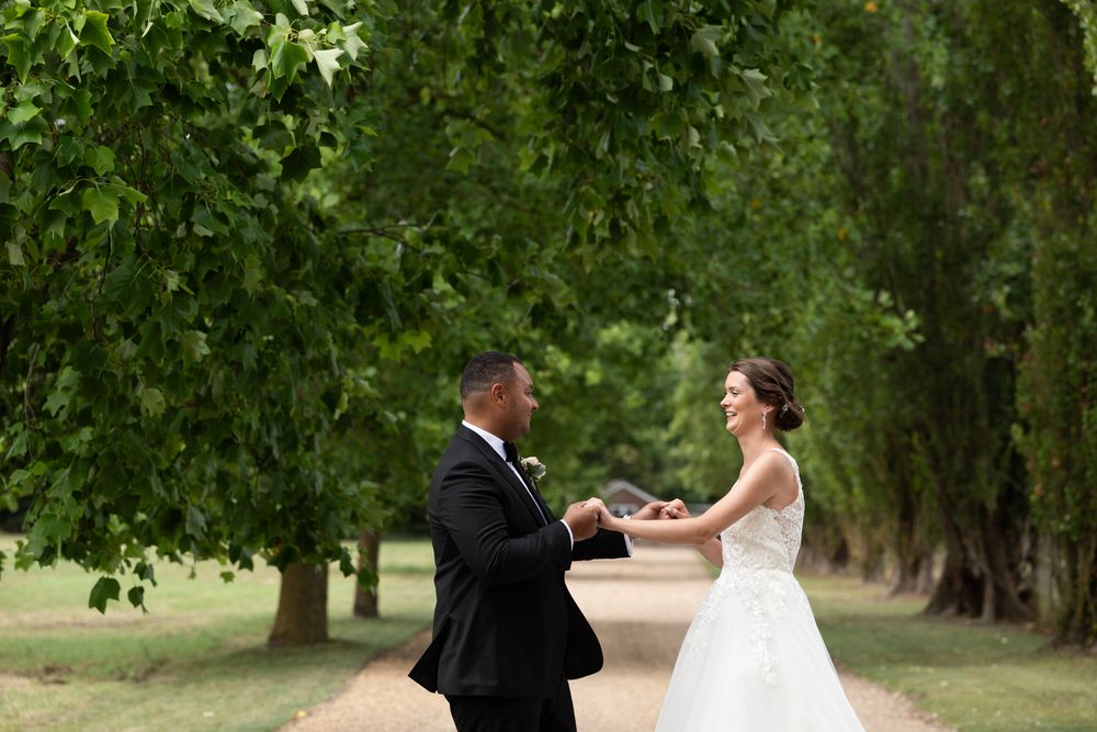 A couple in wedding attire dances together on a lush green lawn surrounded by trees.
