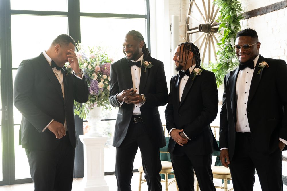 Groomsmen in black tuxedos gathered together in an elegant indoor venue with natural lighting.