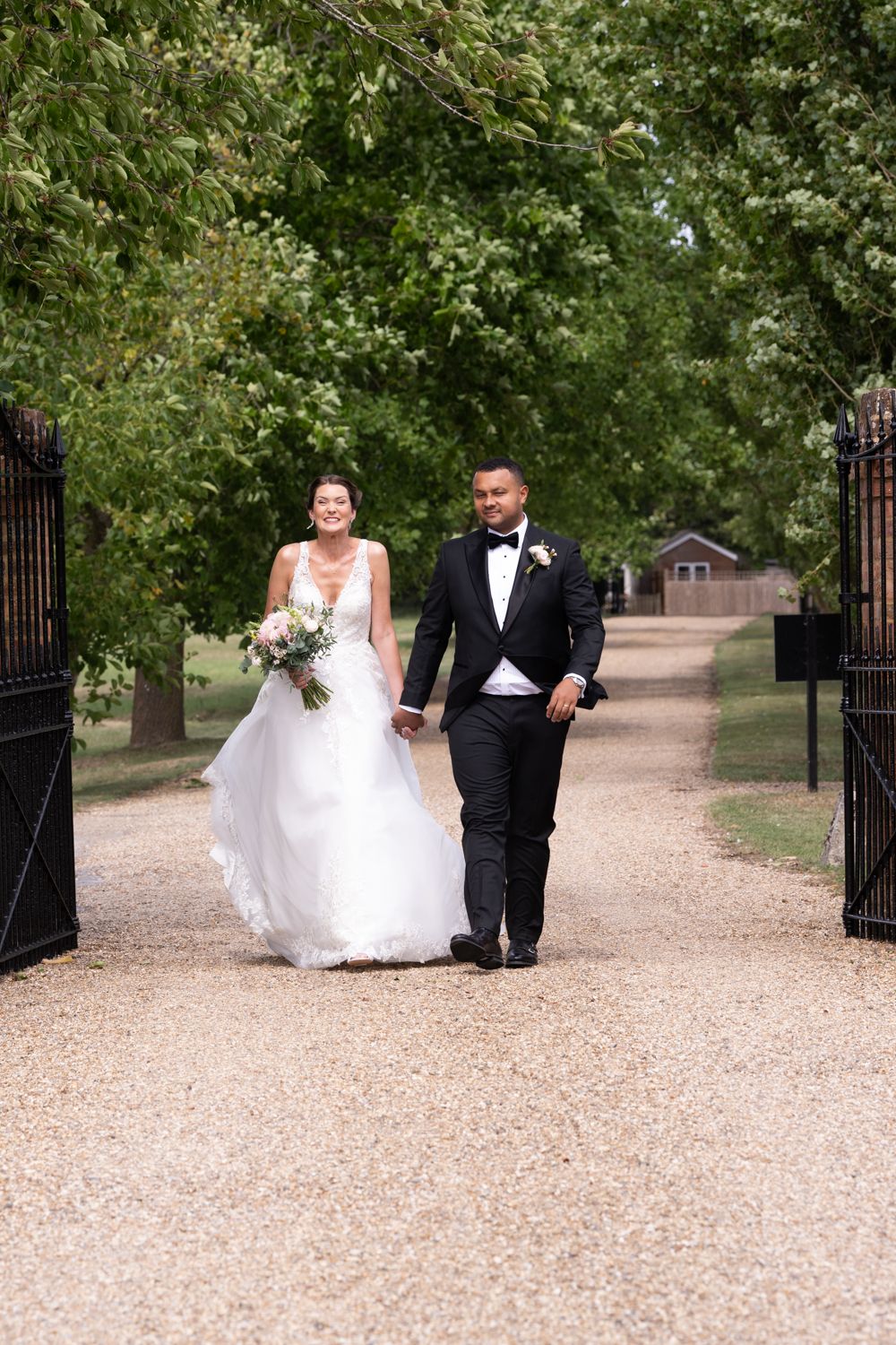 A couple walking together down a tree-lined path through ornate black gates on their wedding day.