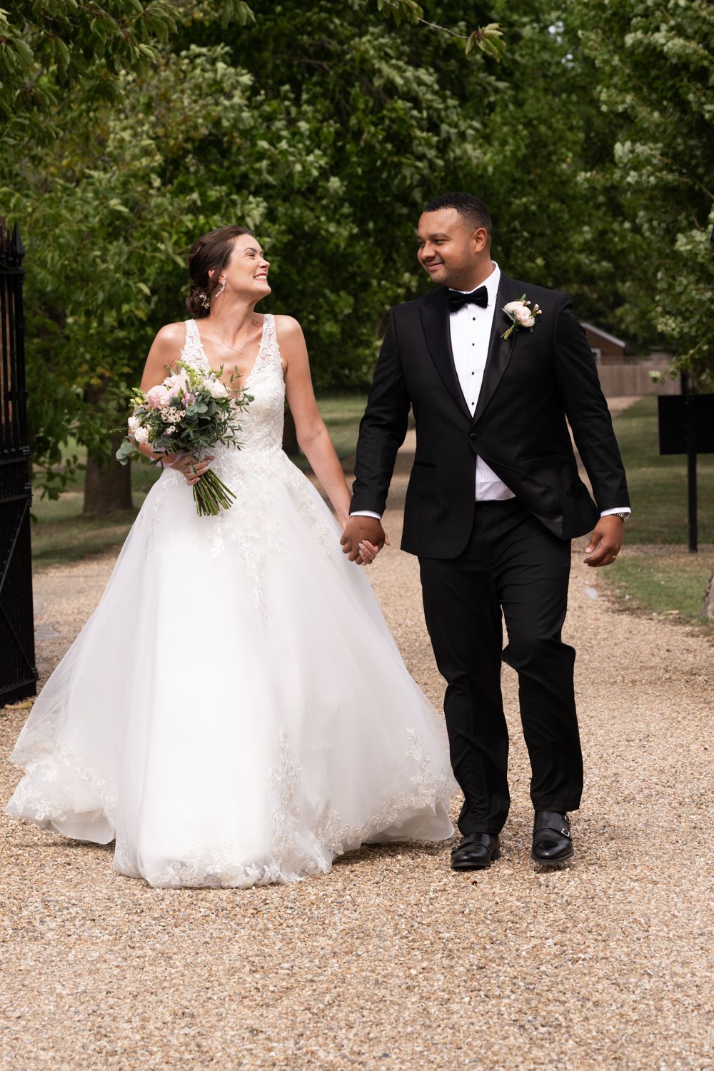 A bride and groom walk hand in hand along a gravel path surrounded by lush green trees.
