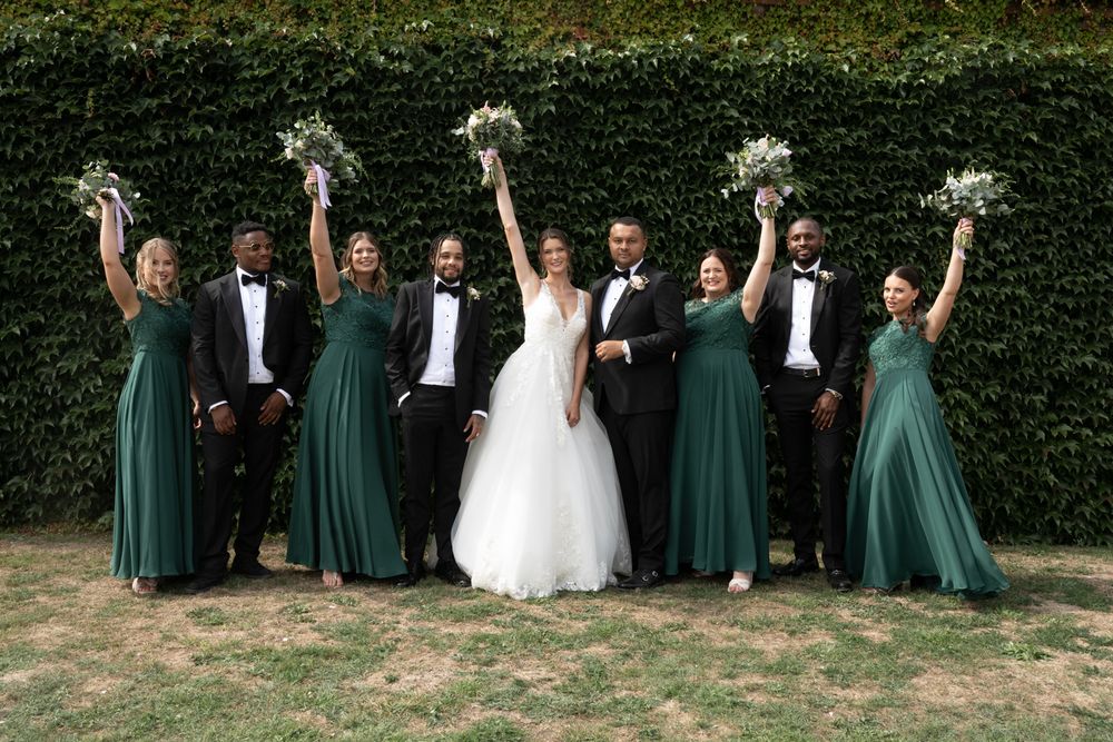 Wedding party in green bridesmaid dresses and black tuxedos poses together outdoors for celebratory group photos.