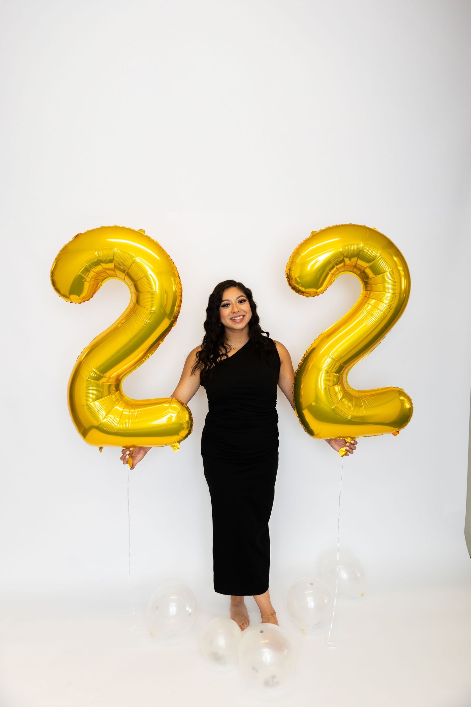 A person in a black dress stands between large gold number 22 balloons against a white background.