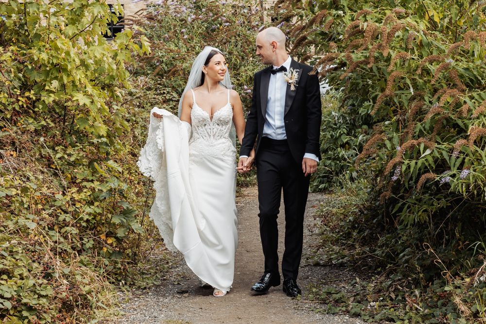 Newlywed couple walks together through lush autumn foliage in a romantic garden setting.