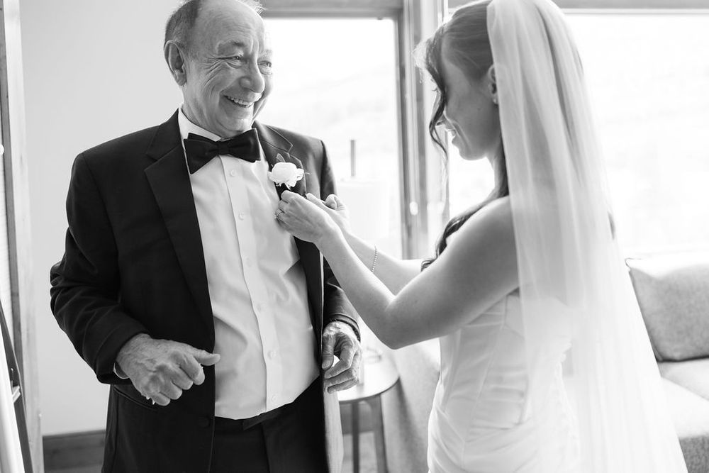 A couple shares a tender moment in formal wedding attire near bright windows.