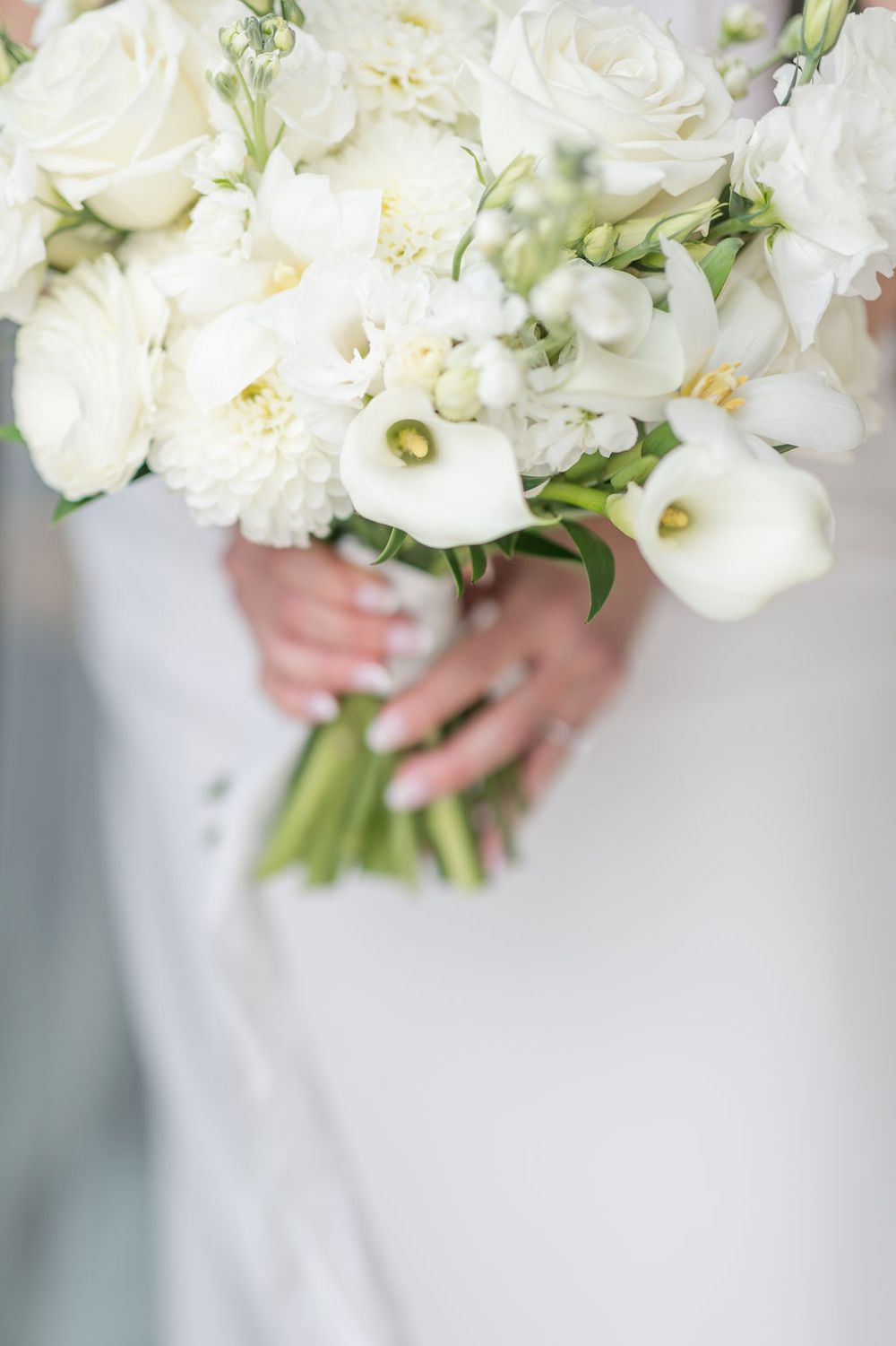Elegant white peony and rose wedding bouquet with delicate greenery held against a light background.