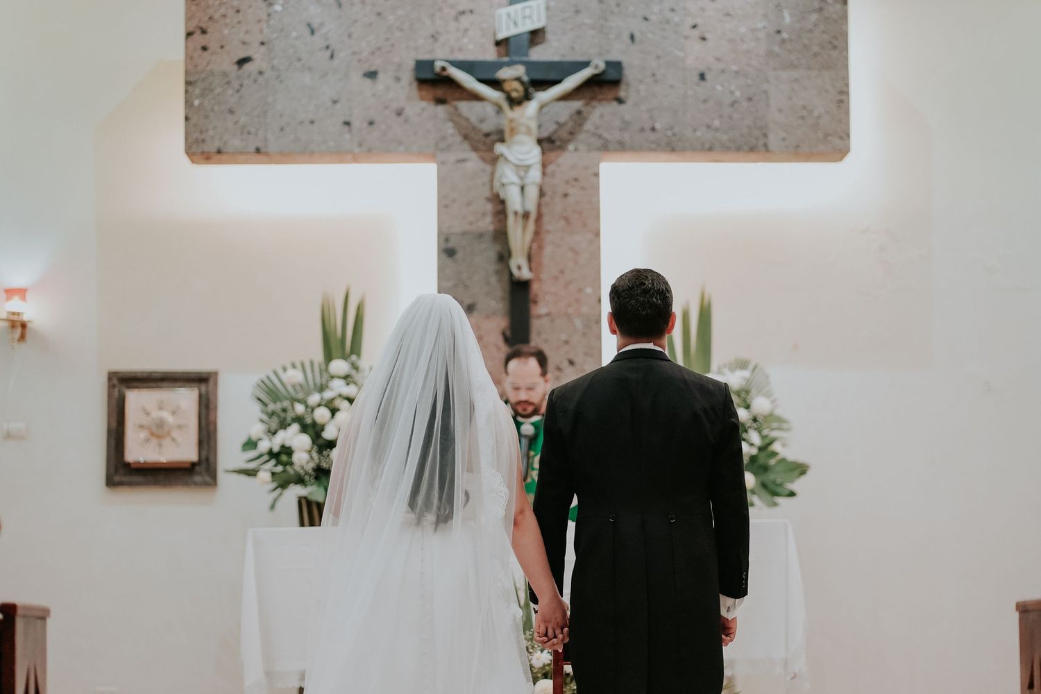 A bride and groom stand at the altar during a Catholic church wedding ceremony with a crucifix above.