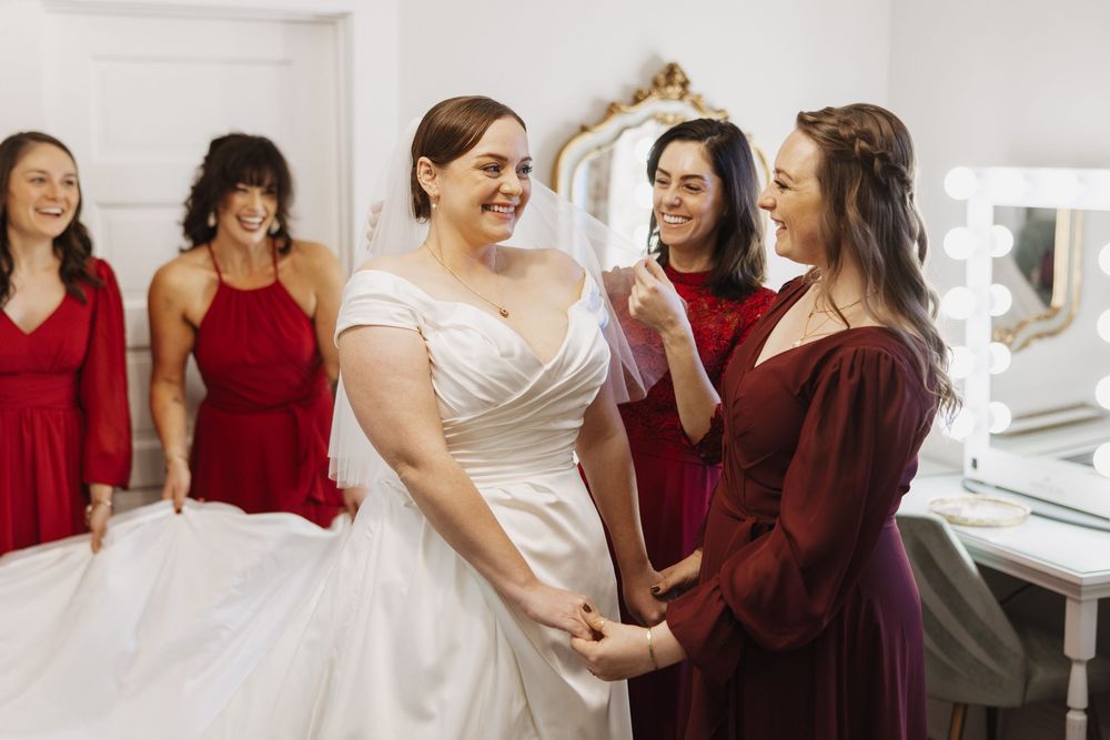 A bride in a white dress stands with bridesmaids wearing burgundy dresses while getting ready in a dressing room.