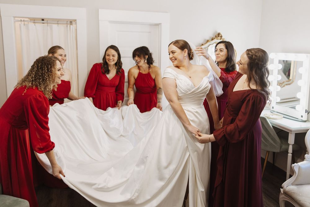 A group of bridesmaids in burgundy dresses help the bride get ready in a bridal suite before the wedding ceremony.