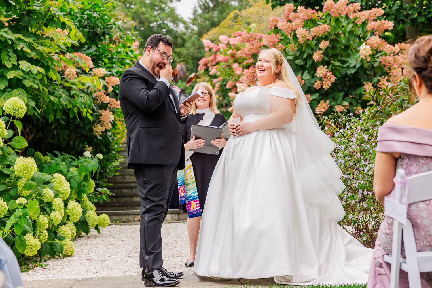 A joyful outdoor wedding ceremony takes place in a lush garden with blooming hydrangeas and pink roses.