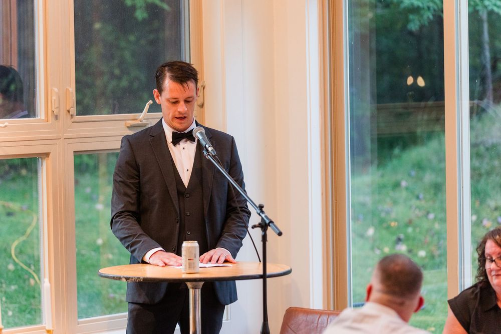 A speaker delivers a speech at a wedding reception in an elegant room with large windows and natural lighting.