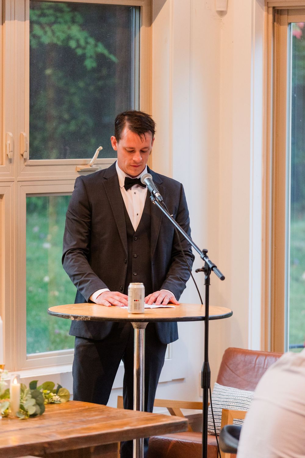 A person in a formal black tuxedo and bow tie stands at a podium giving a speech during an indoor event.