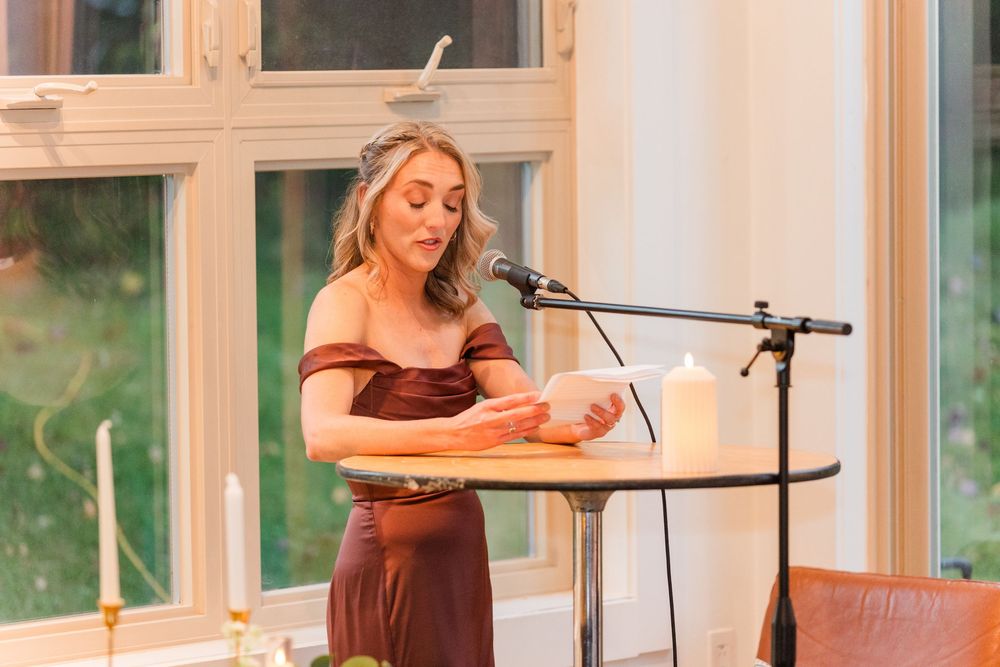 Someone in a brown dress reads from a podium at a microphone stand near large windows.