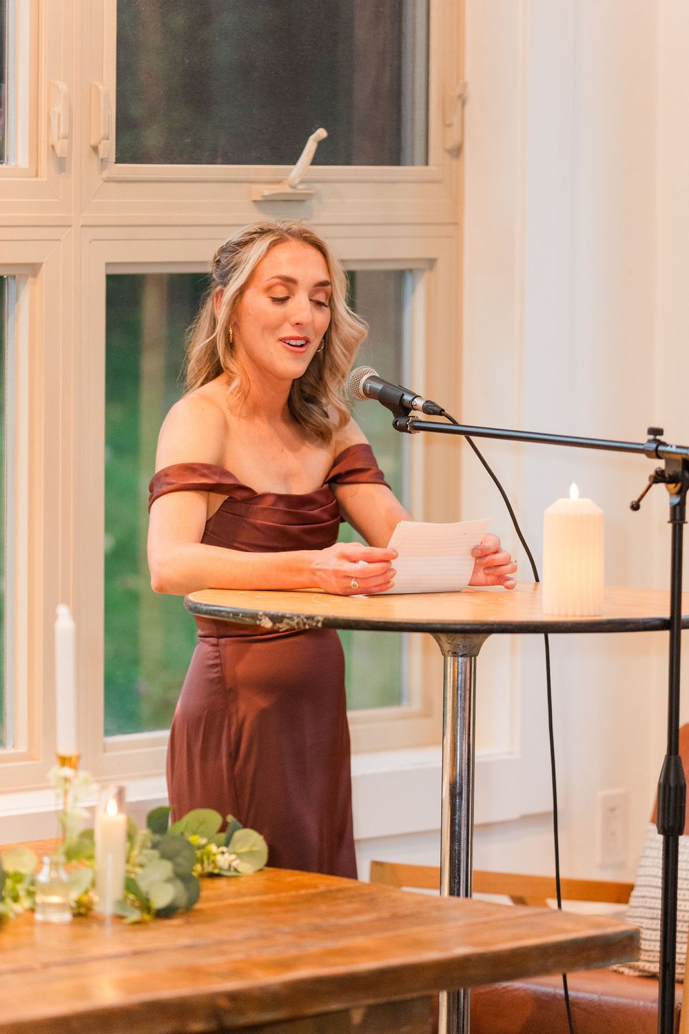 Wedding guest in burgundy dress reads speech from paper at wooden table during reception celebration.