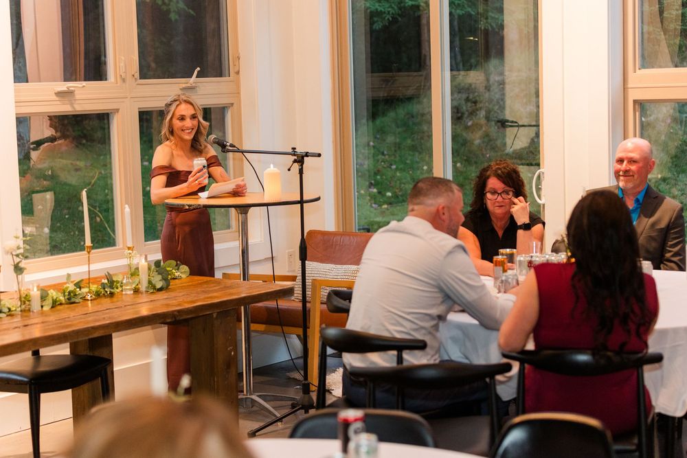 People gather around long wooden tables in a bright room with large windows during a social event.