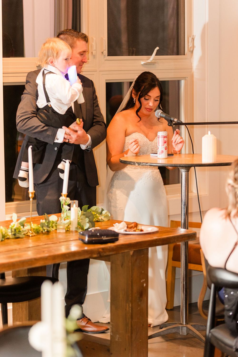 Wedding reception guests enjoy dinner and conversation at long wooden tables with white tablecloths and candlelight.