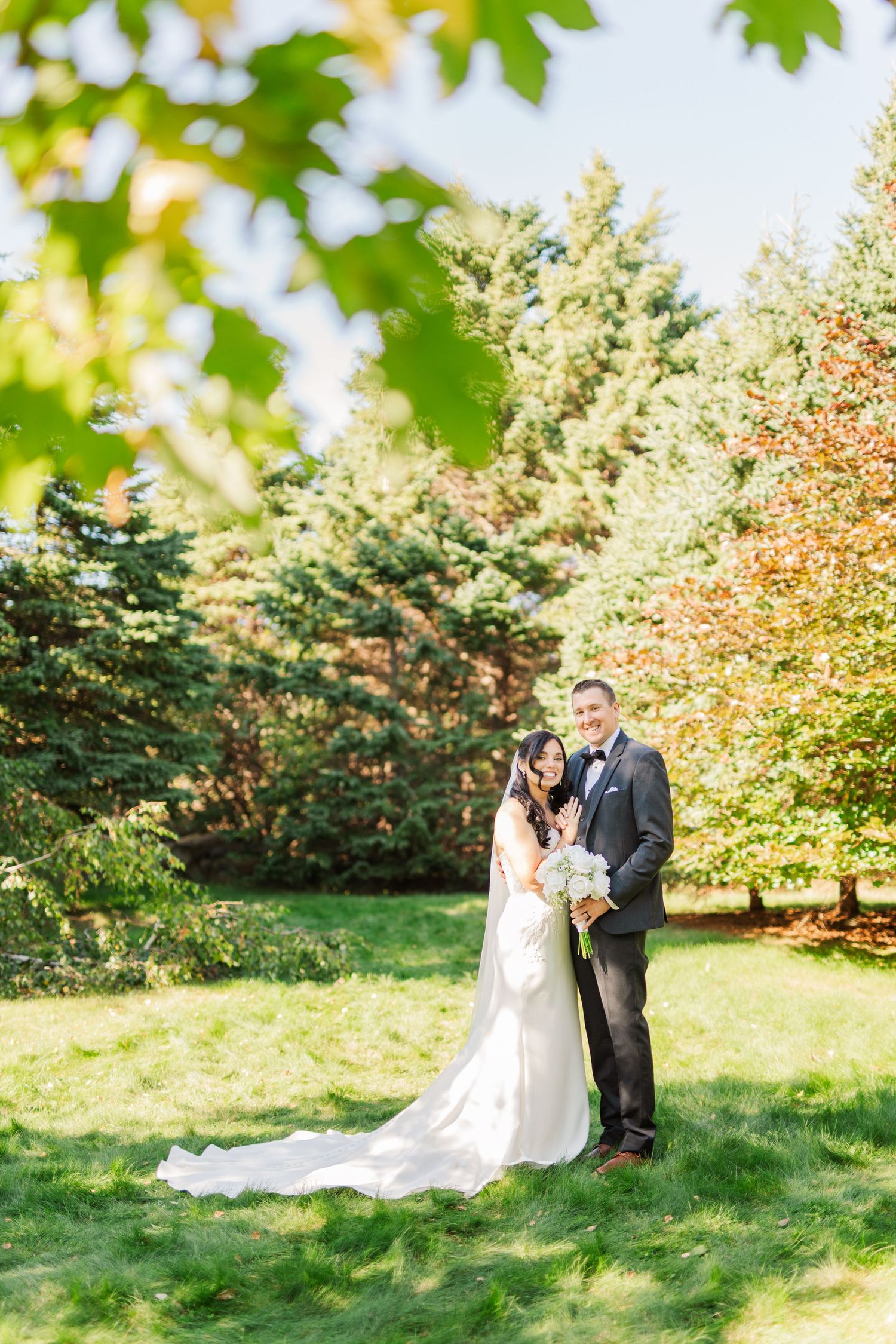 A couple in wedding attire embrace on a grassy hill surrounded by autumn trees with changing leaves.