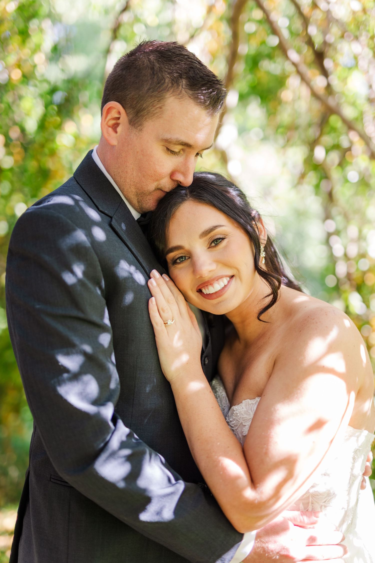 A romantic wedding portrait outdoors with dappled sunlight filtering through trees onto the happy couple.