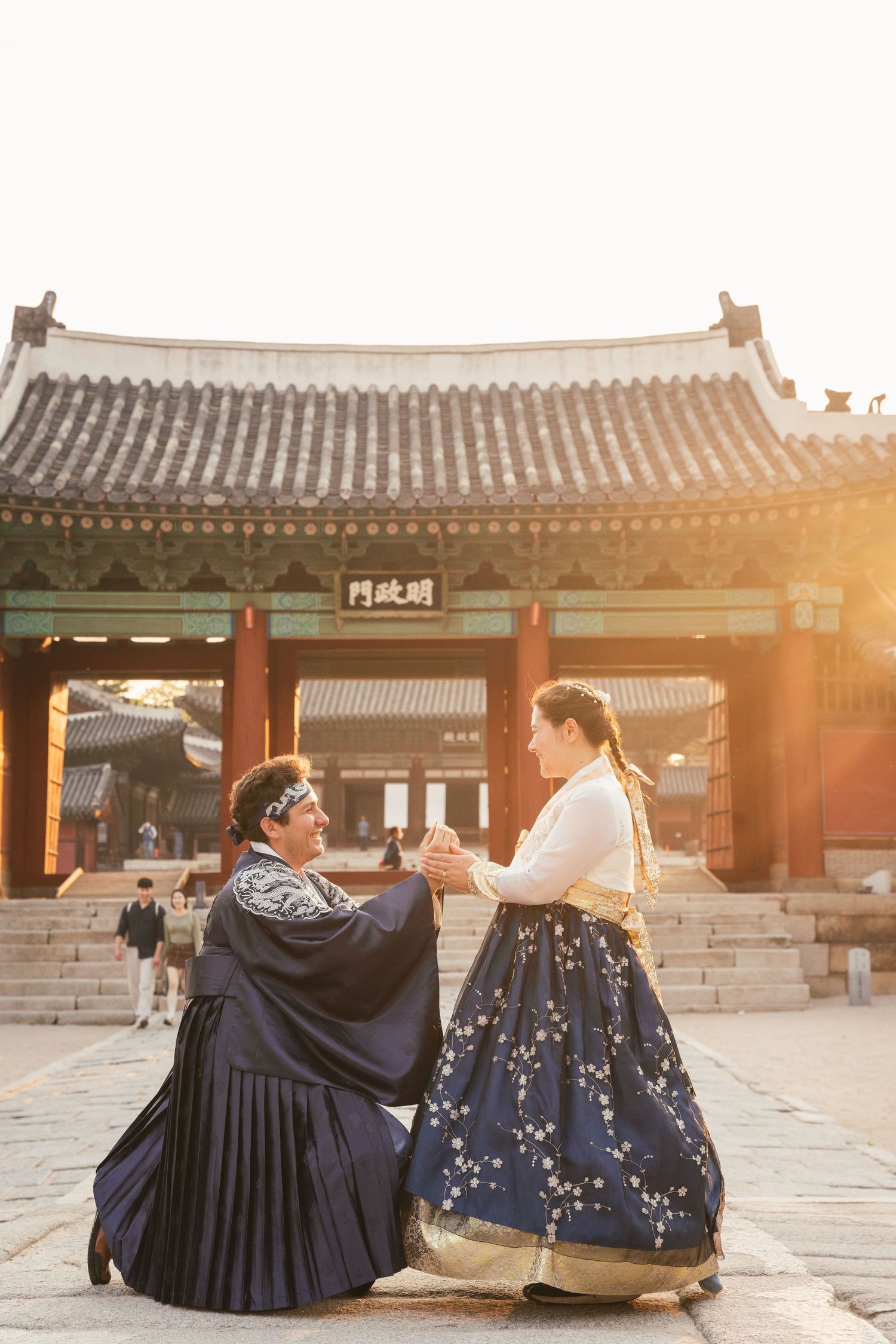 Two people in traditional Korean hanbok dresses sit together at a historic palace courtyard during golden sunset.