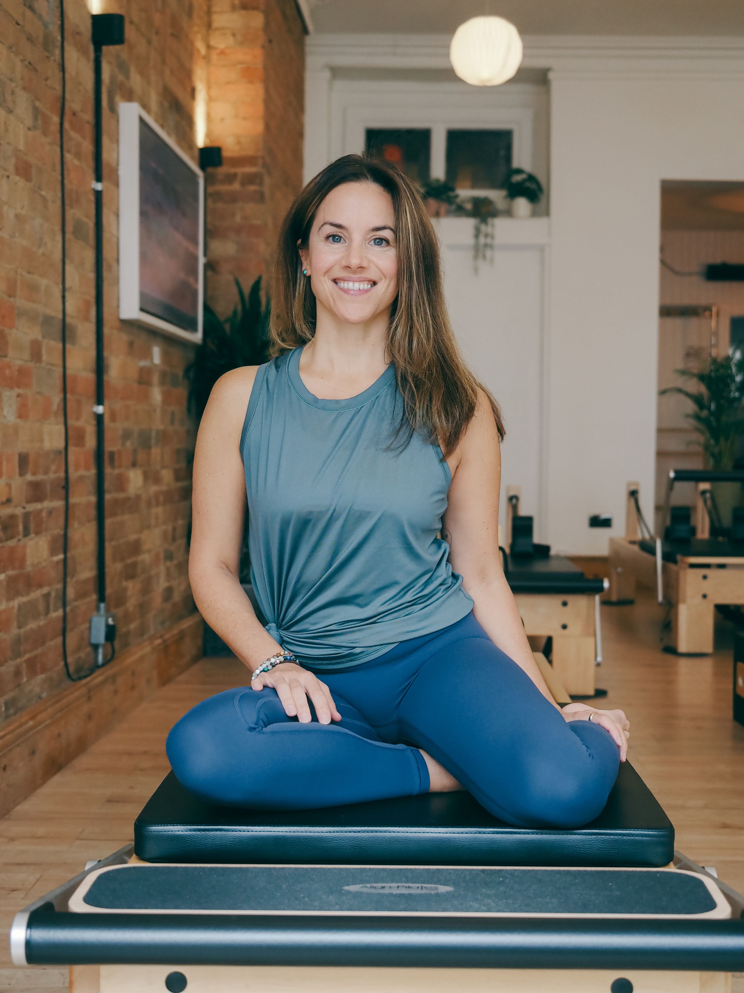 Fitness instructor sitting cross-legged on a Pilates reformer machine in a studio with exposed brick walls.