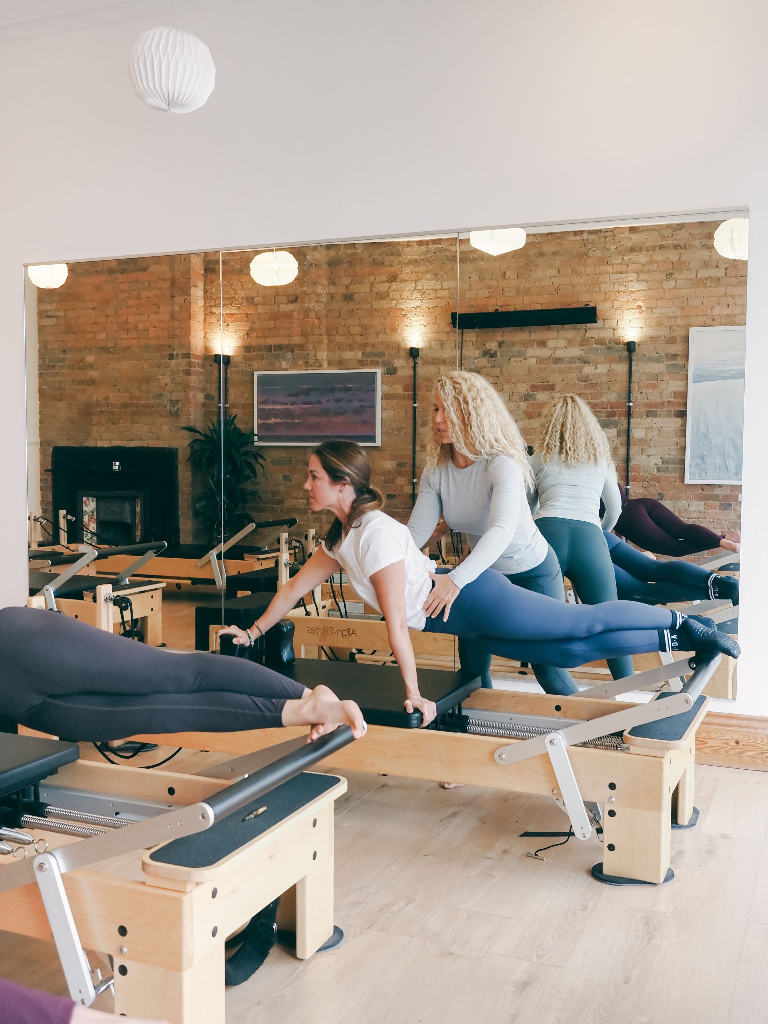 Three people practicing Pilates exercises on reformer machines in a bright studio with exposed brick walls.