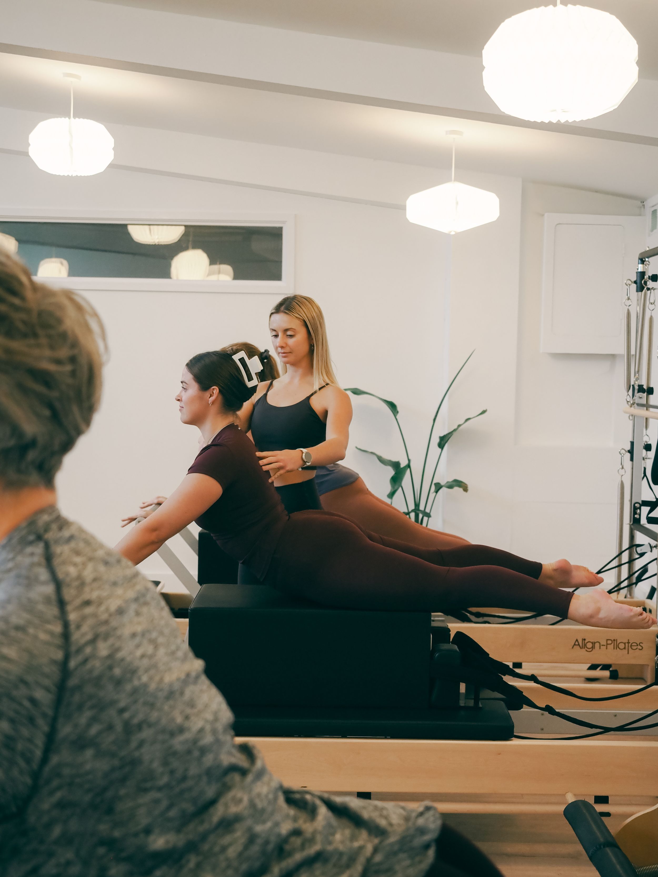 Pilates students practicing exercises on reformer machines in a bright studio with white pendant lights.