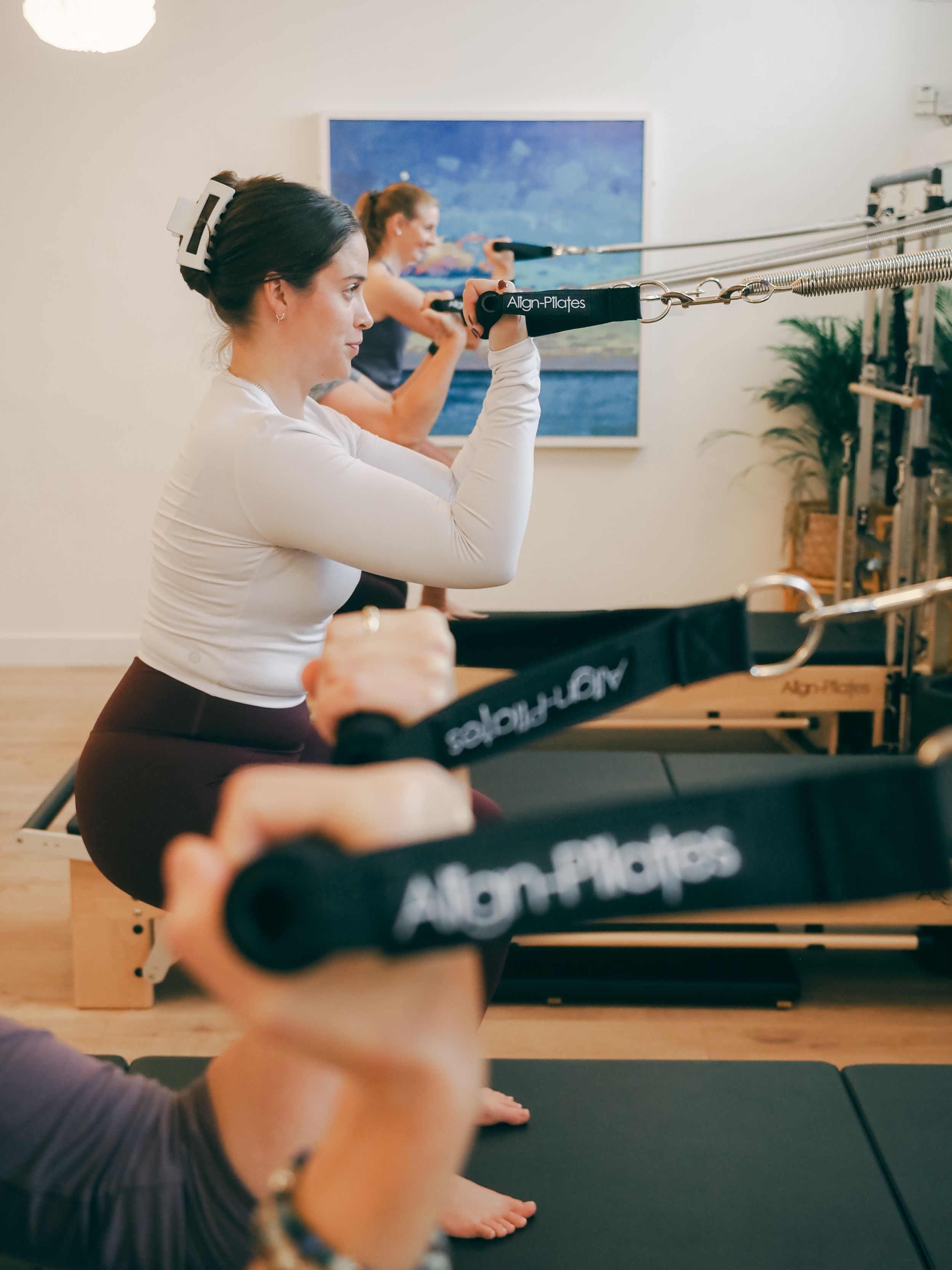 People exercising on Pilates reformer machines in a bright studio with blue artwork on the wall.