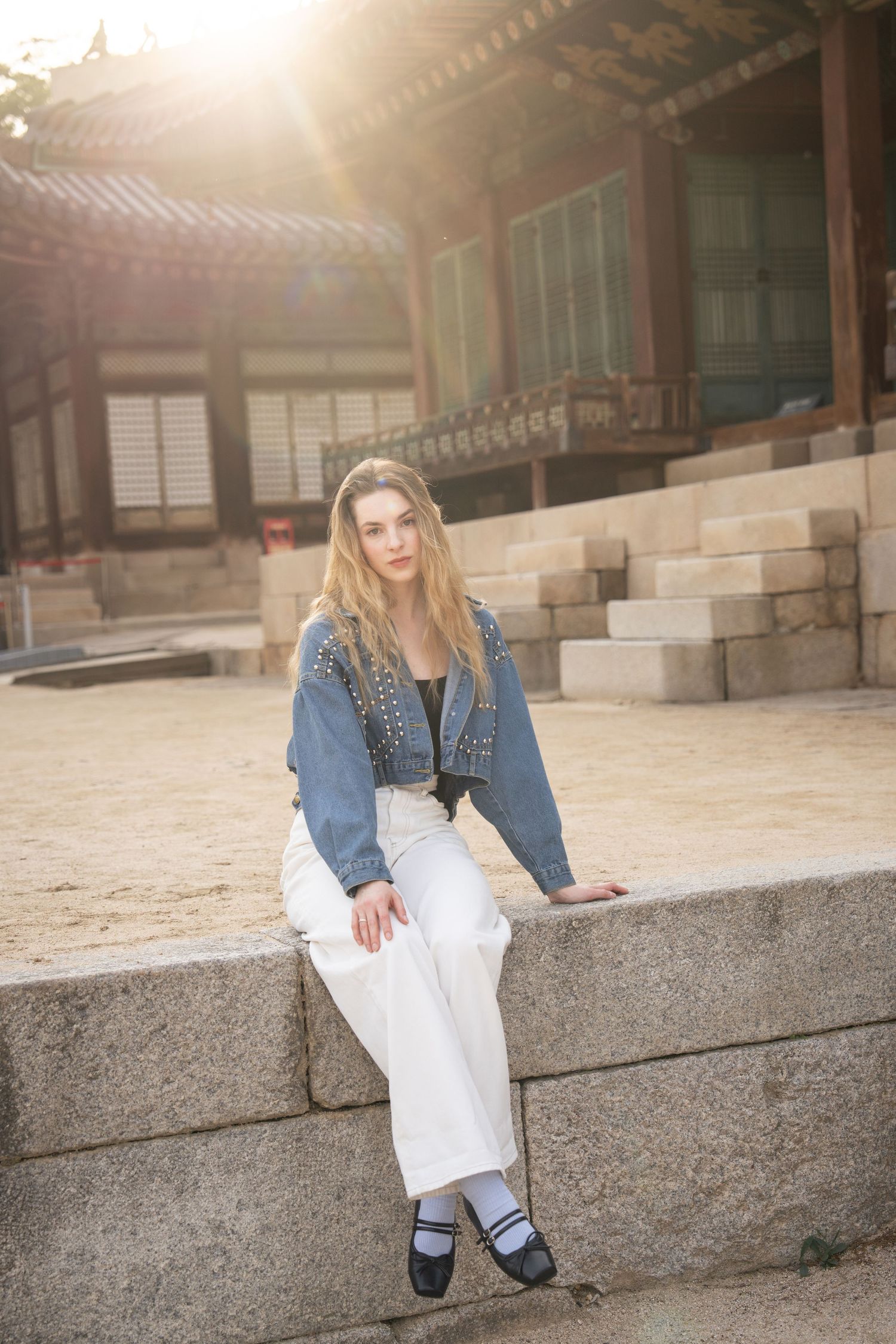 Person in denim jacket and white pants sits on stone steps at traditional Asian temple courtyard in warm sunlight.