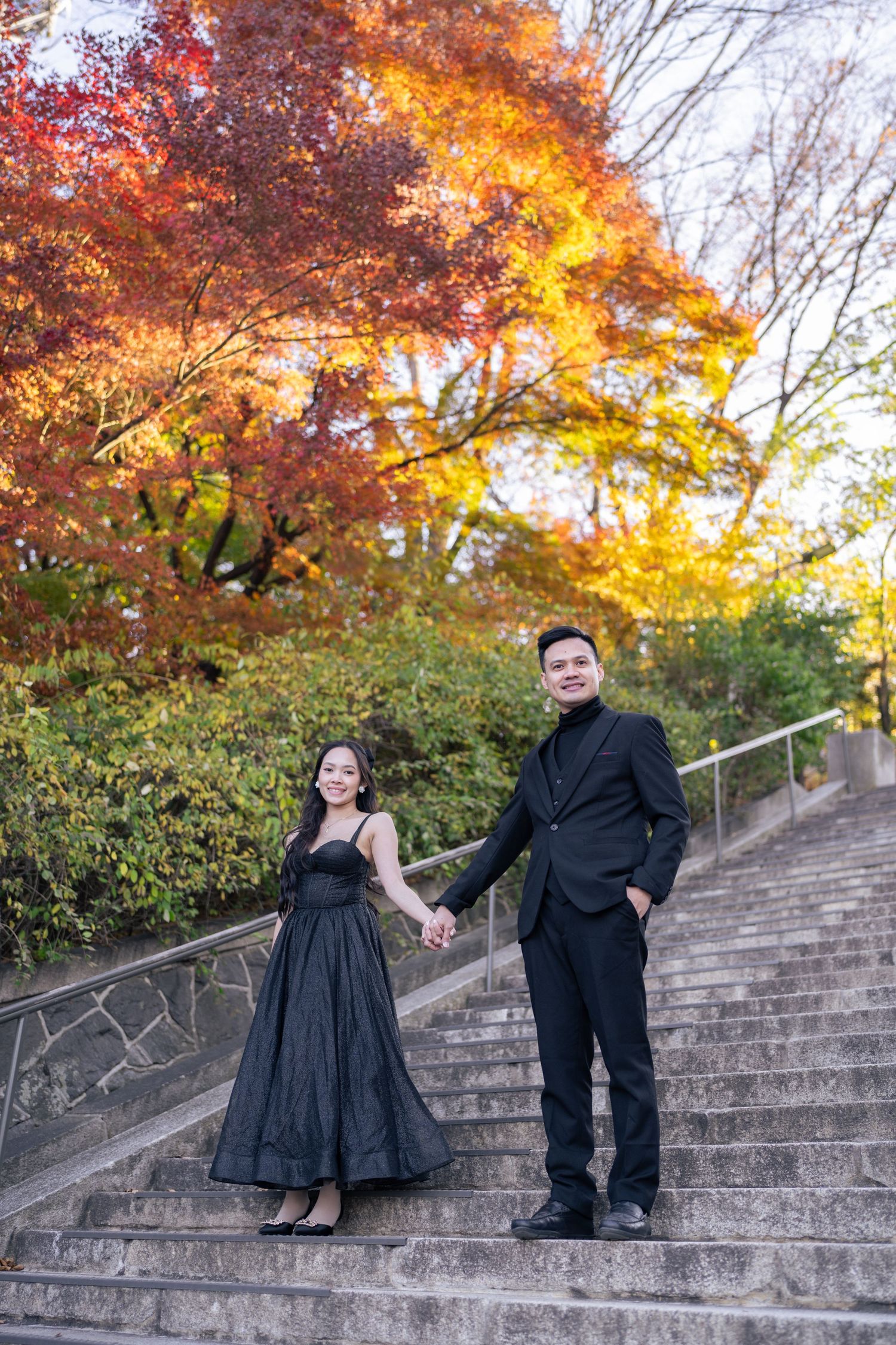A couple poses playfully on concrete steps surrounded by vibrant autumn foliage with orange and red maple leaves.