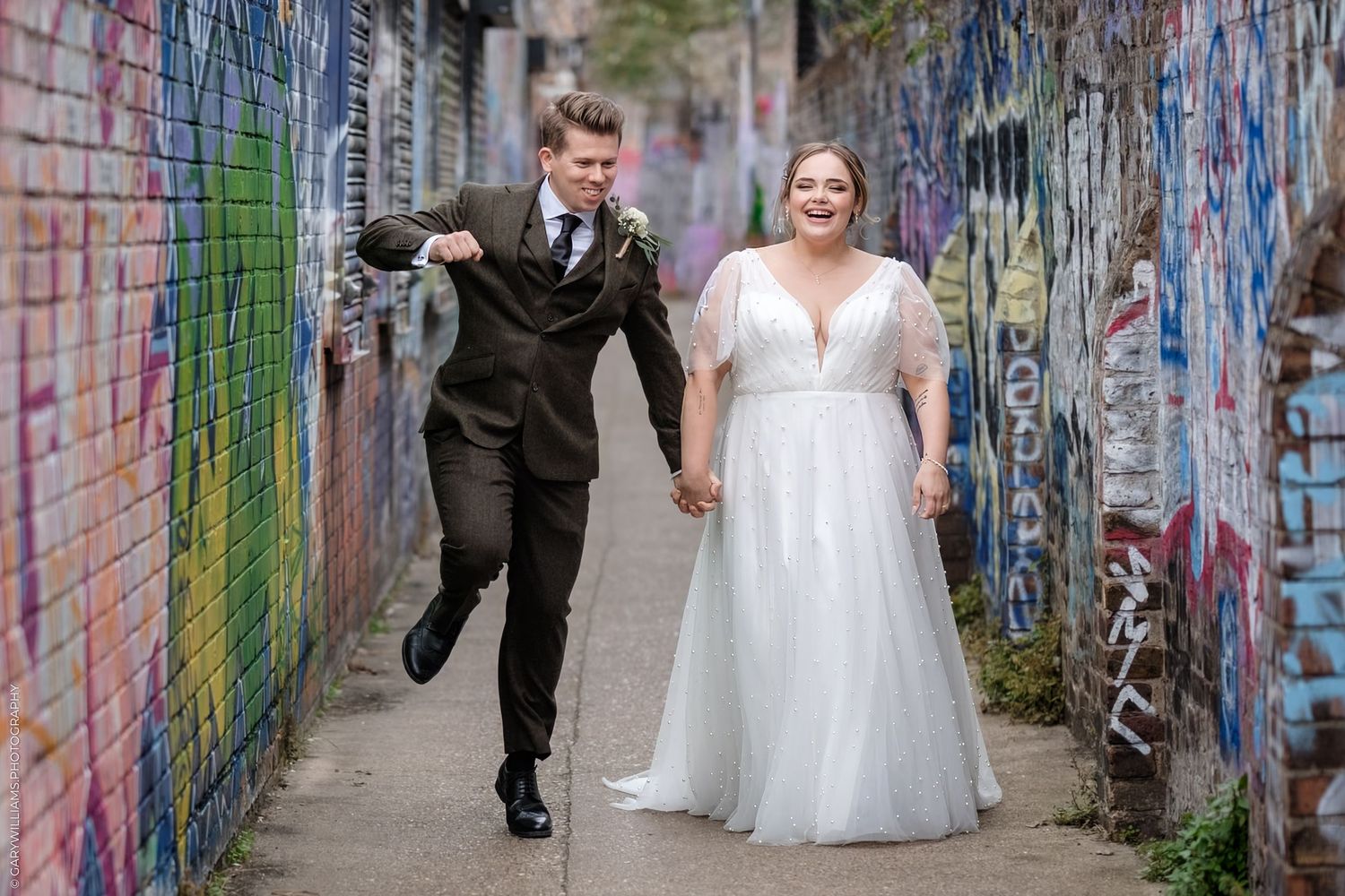 Wedding couple walks along colorful graffiti wall in urban alleyway while photographer captures their special moment.
