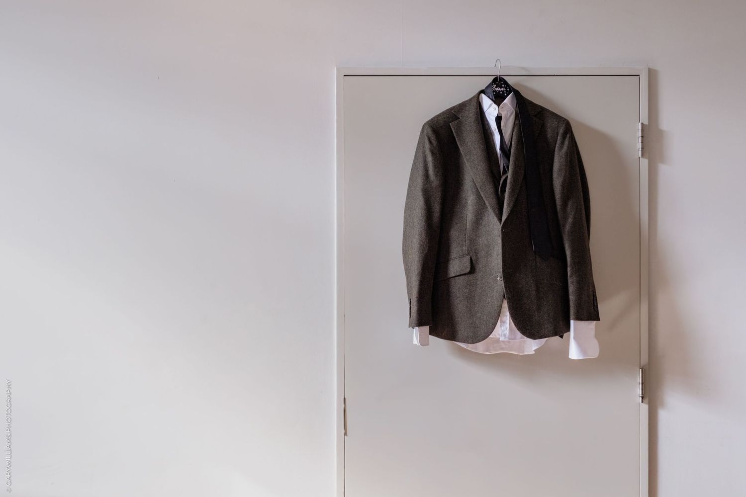 A row of formal suits and dress shirts hanging neatly on white hangers against a light colored wall.