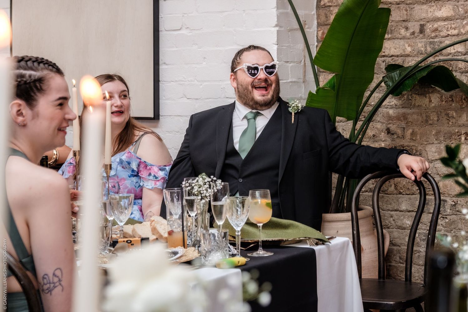 Wedding celebration scene with tables decorated with greenery and warm lighting in an industrial-style venue.