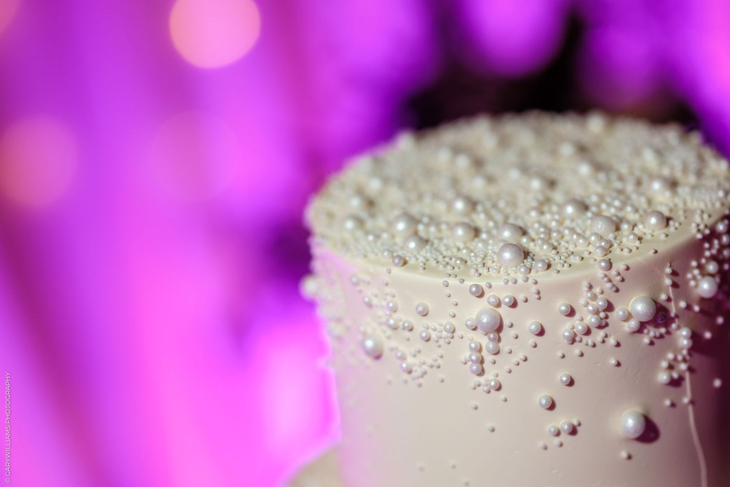 Close-up shot of a white frosted cake with pearl sprinkles against a blurred purple bokeh background.