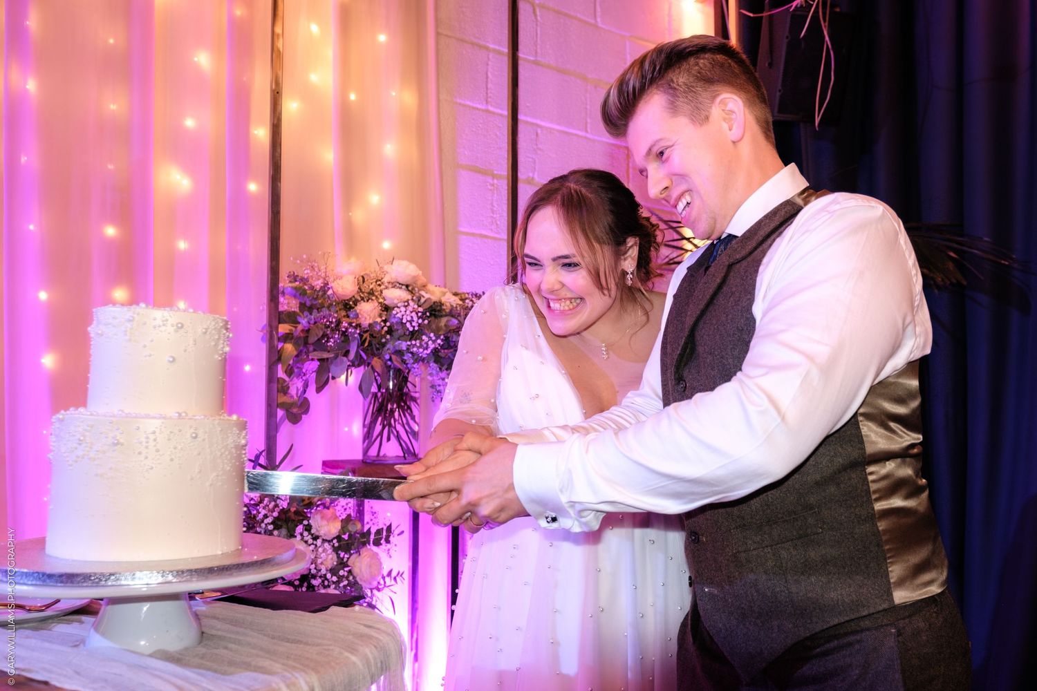 A newlywed couple cuts their elegant white wedding cake together at their pink-lit reception celebration.