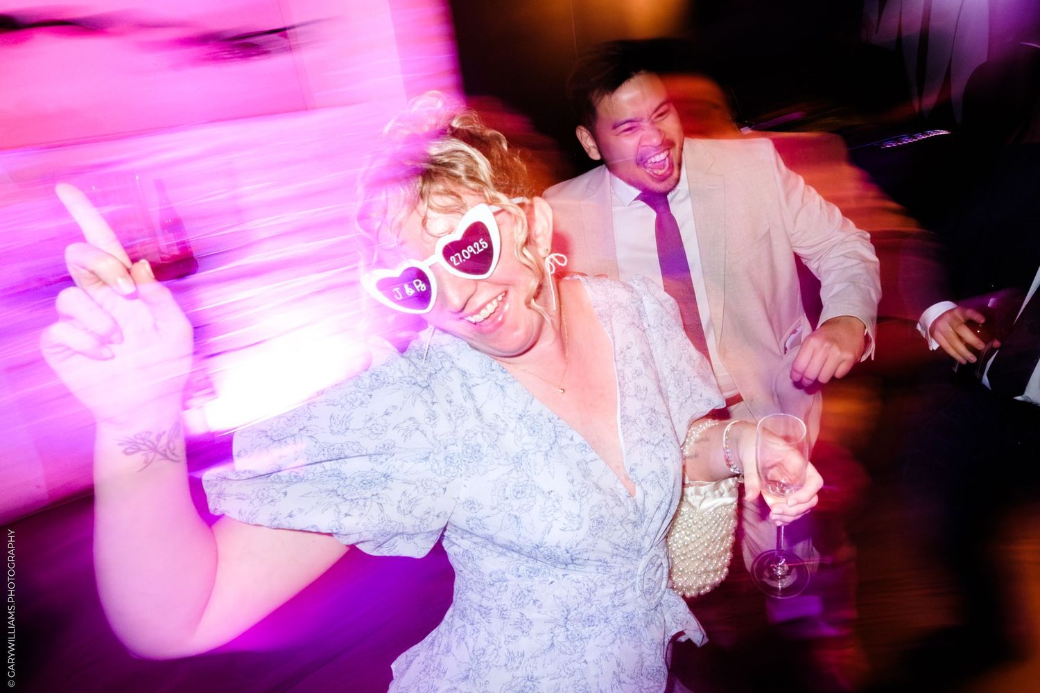 Wedding guests dance and celebrate on a colorful dance floor illuminated by vibrant pink lighting at a reception party.