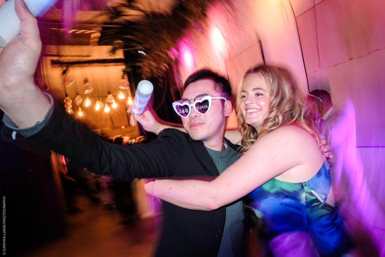 People dancing and celebrating at a lively party with colorful pink and purple lighting effects.