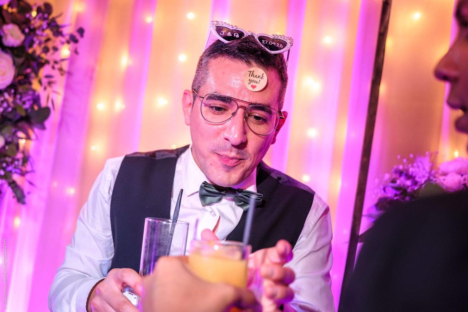 A bartender in a bowtie and vest mixes drinks behind a bar with pink and purple lighting in the background.