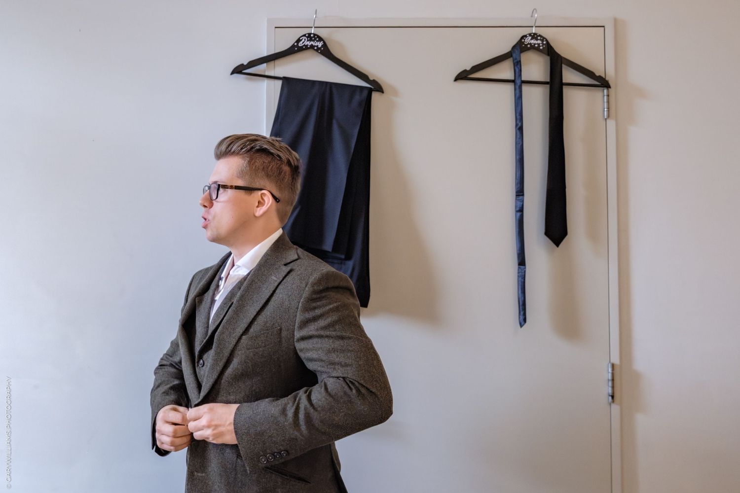 Three views of someone wearing a grey suit and tie standing against a white wall with black graduation gowns hanging behind.