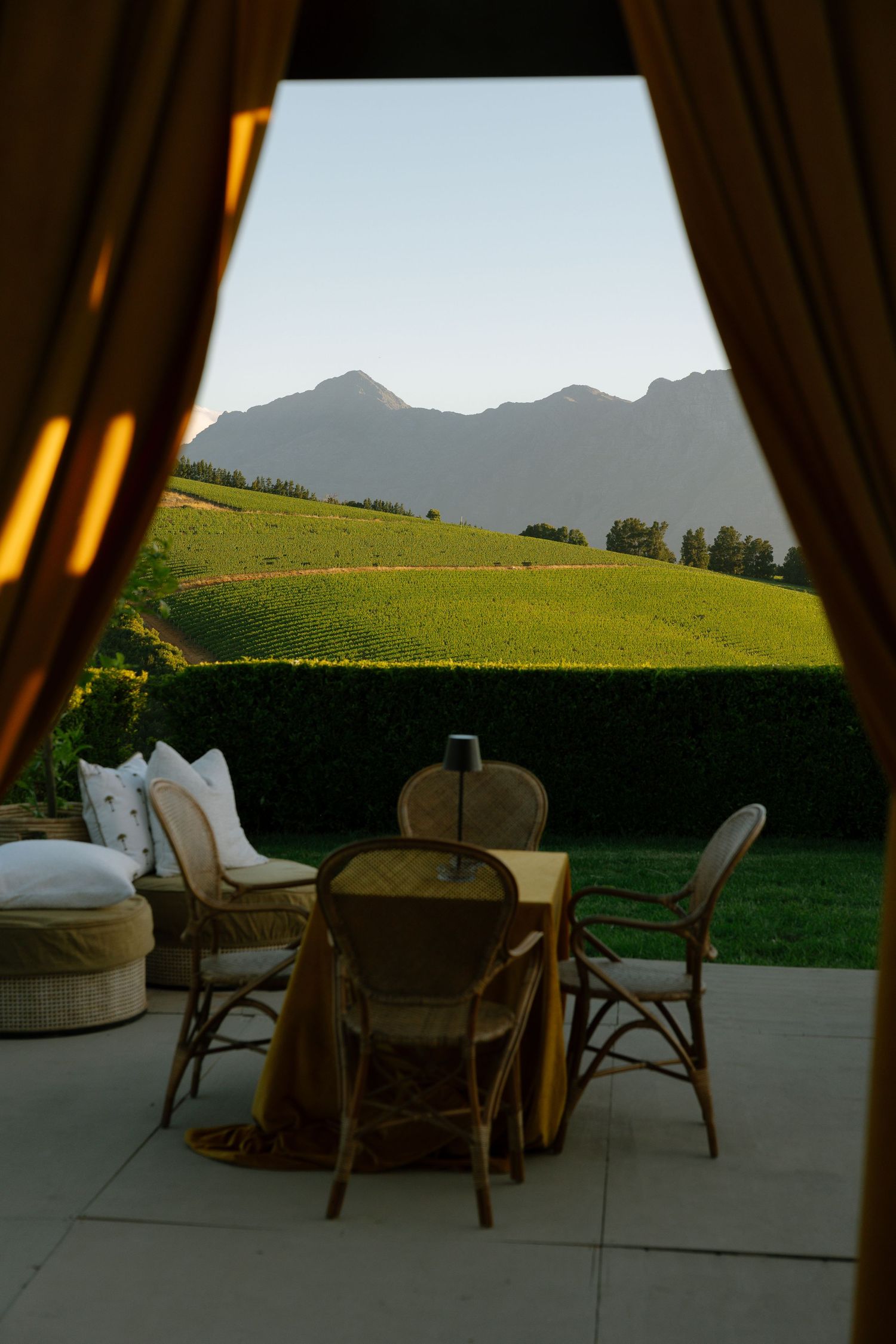 Mountain view through curtains with outdoor seating area featuring rattan chairs and table on a patio overlooking green hills.
