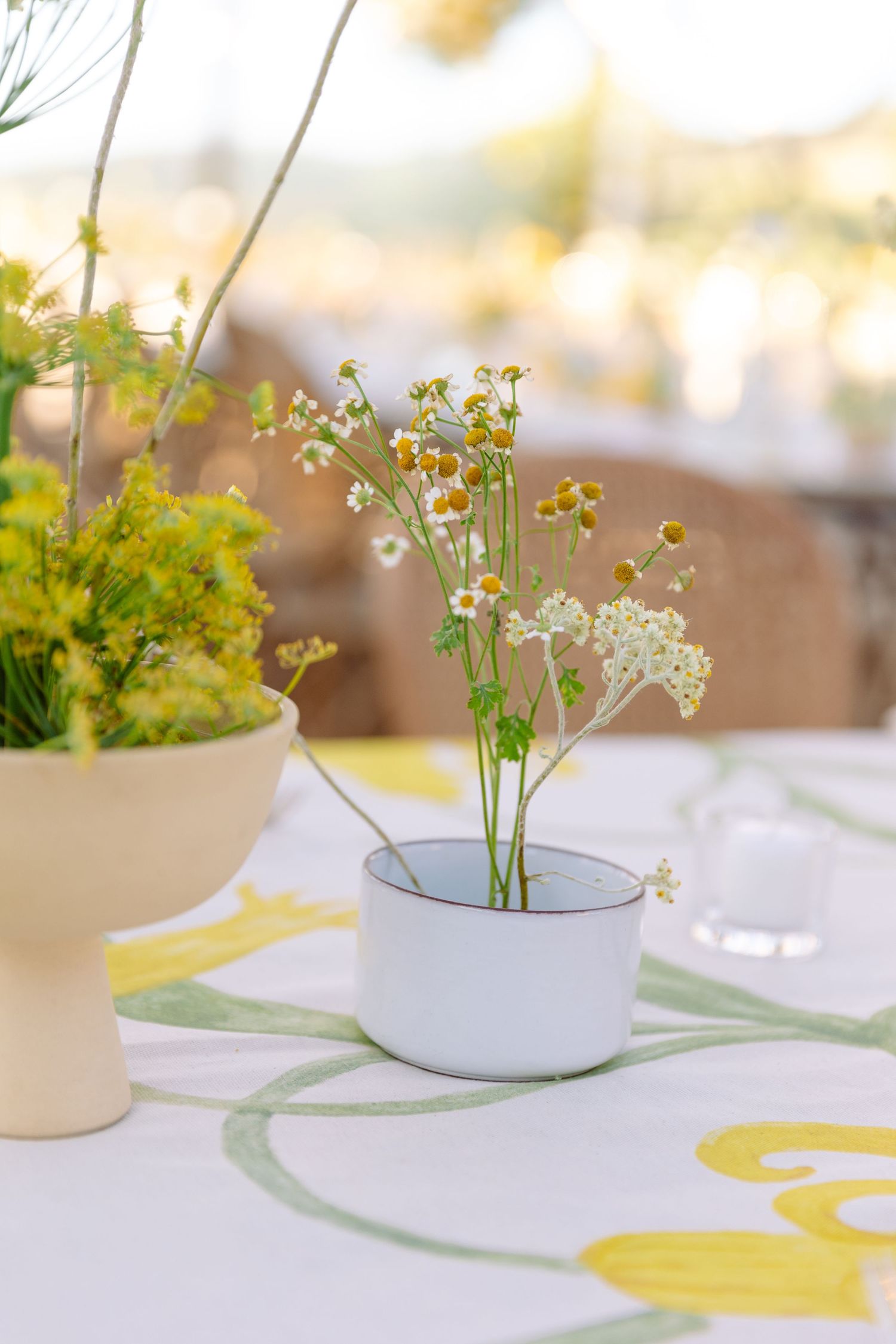 Small delicate white wildflowers in a ceramic cup sit on a table with floral pattern tablecloth in soft natural light.