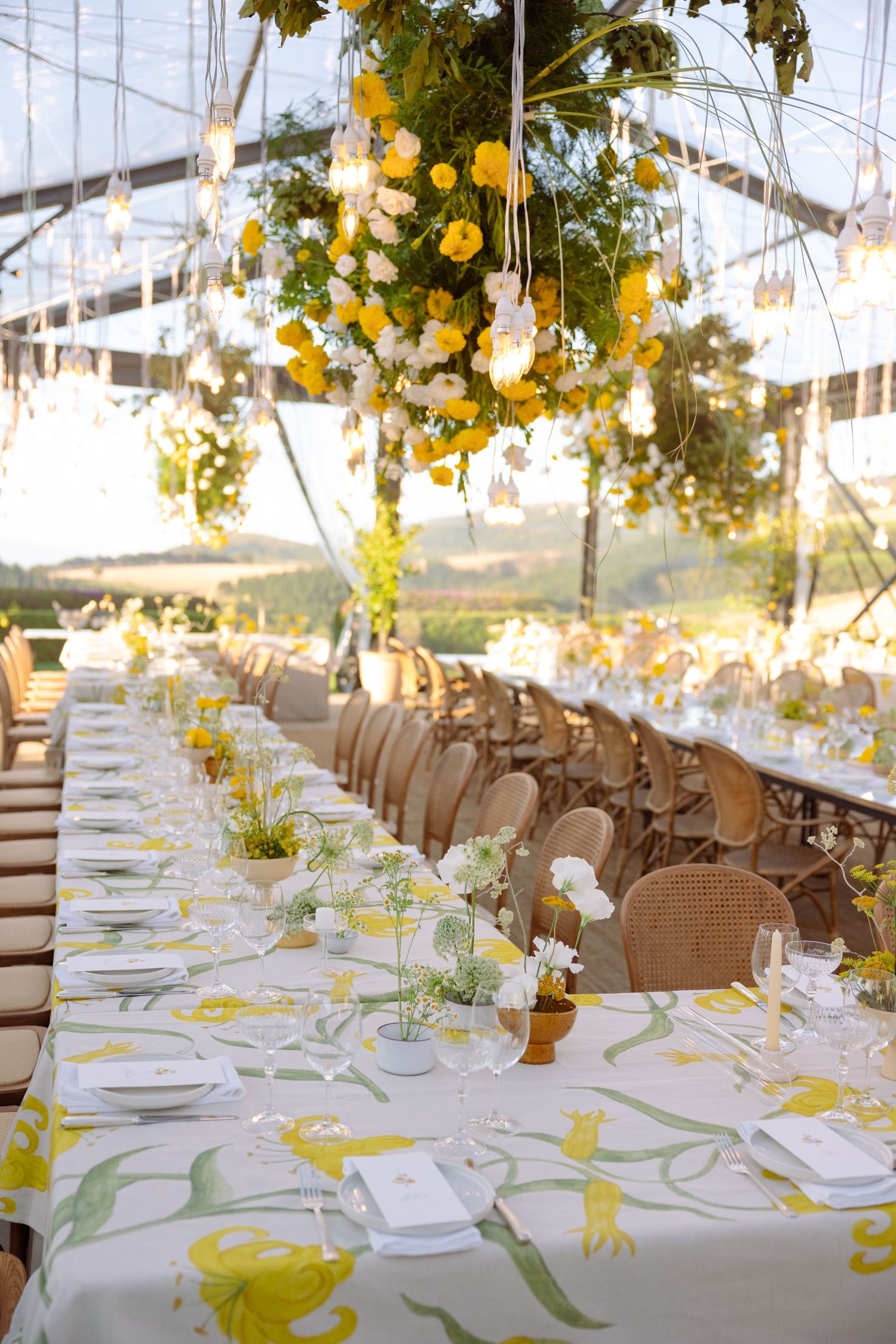 Elegant wedding reception tables set with white linens, yellow floral centerpieces, and place settings under a tent canopy.