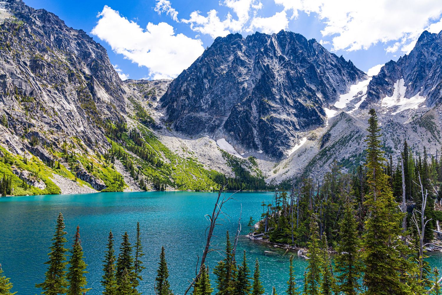 Vivid blue lake with trees on the shoreline and Rocky Mountain in the background under sunny skies.