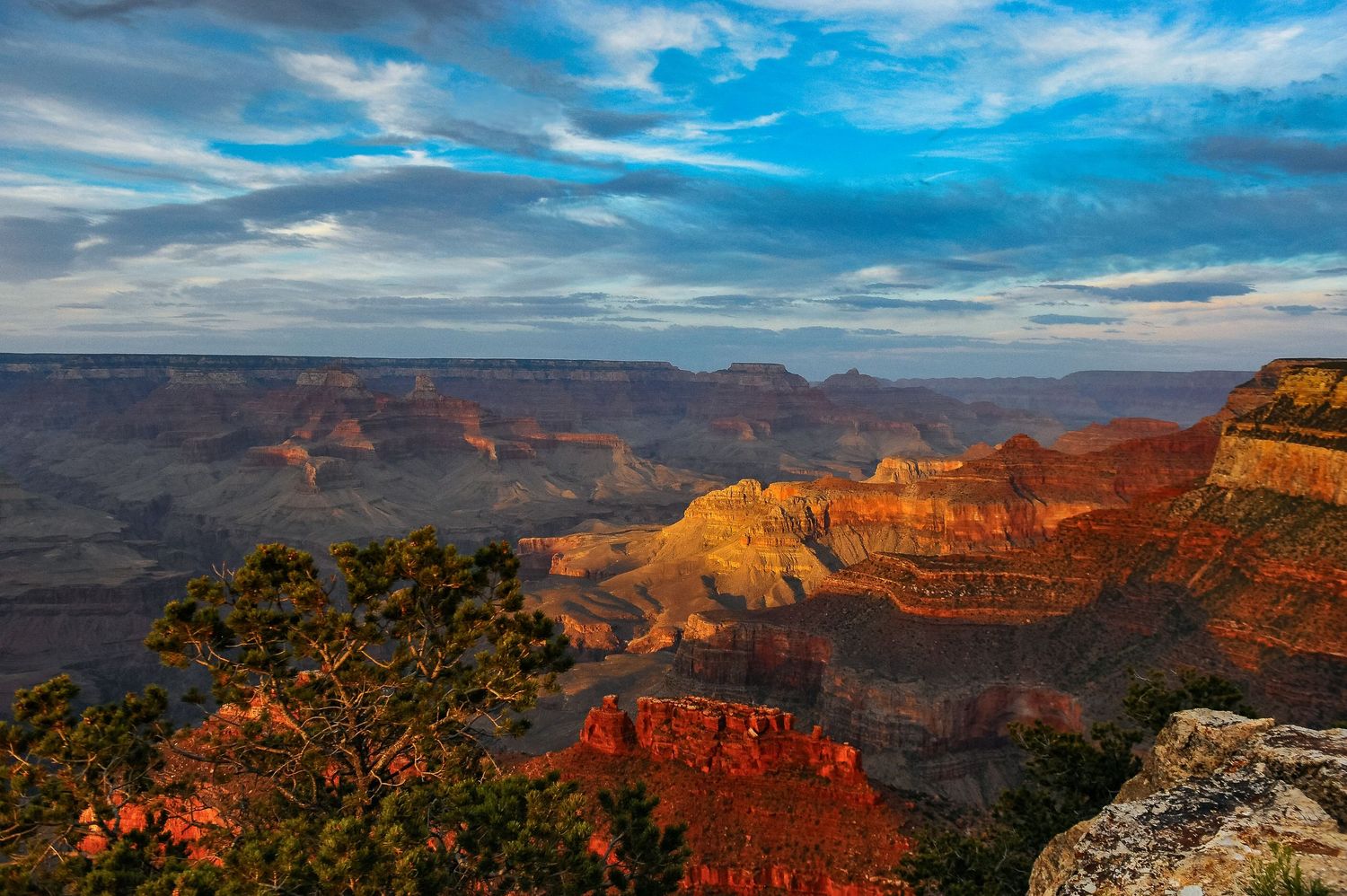 Panoramic view of the Grand Canyon at sunset with vibrant orange cliffs and dramatic blue sky with scattered clouds.