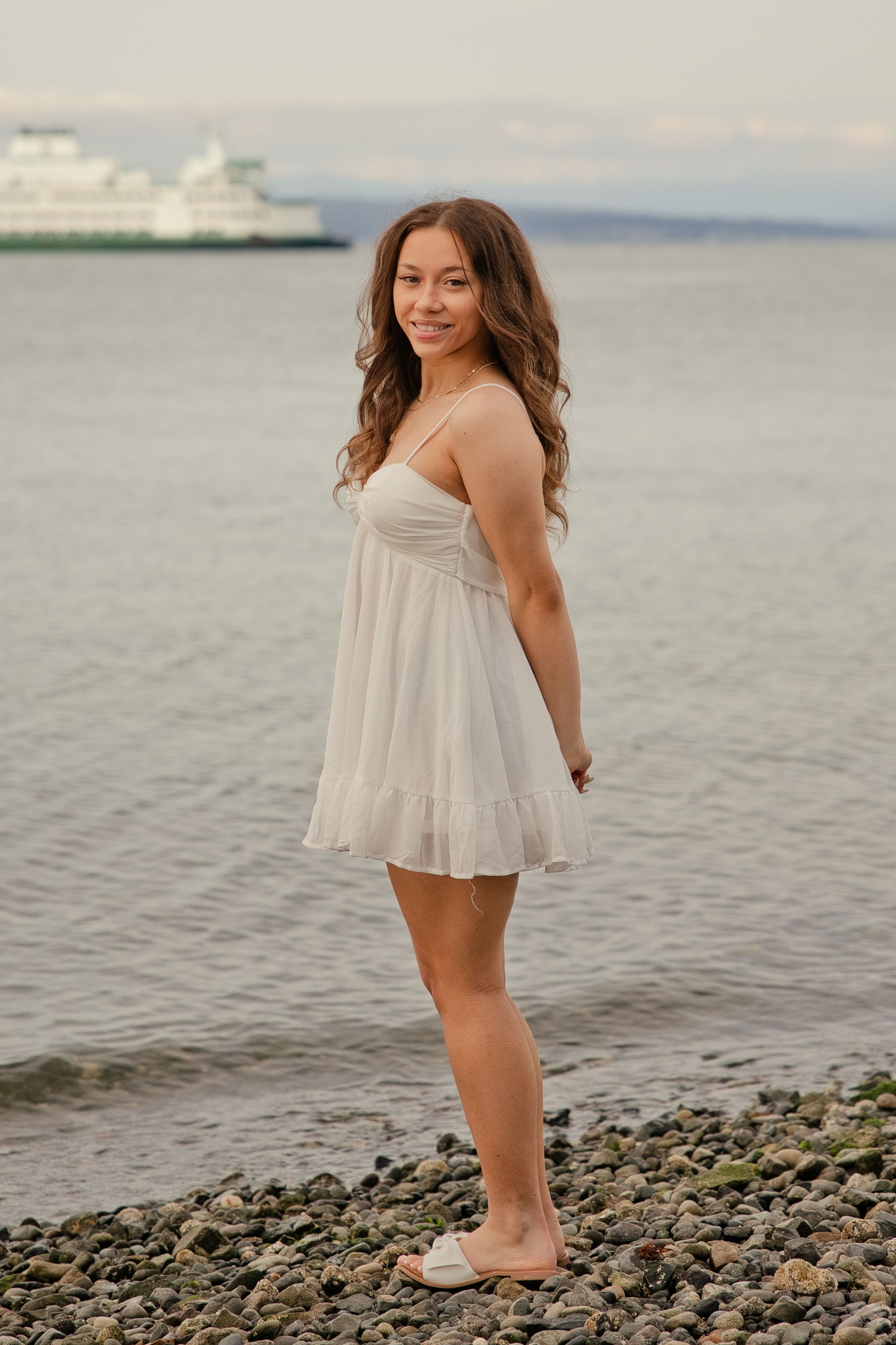 Young woman in white sundress standing on rocky beach shore with calm water and ferry boat in background.
