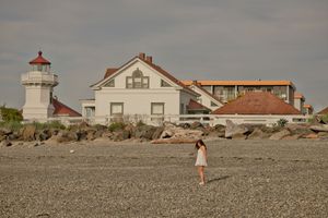 Woman in white dress walking on rocky beach near historic lighthouse and white coastal building.