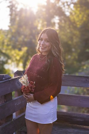 A person in a burgundy sweater and white top poses against a wooden railing at sunset with golden light.