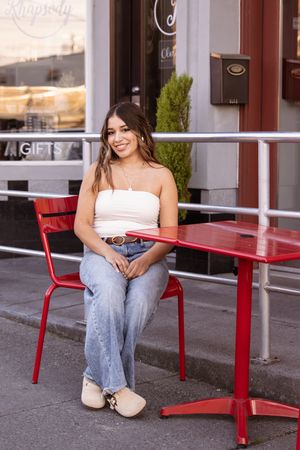 Person in jeans sits on red metal bench outside storefront with 'GIFTS' signage visible.