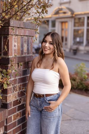 Woman in white crop top and high-waisted jeans poses against brick wall in urban setting.