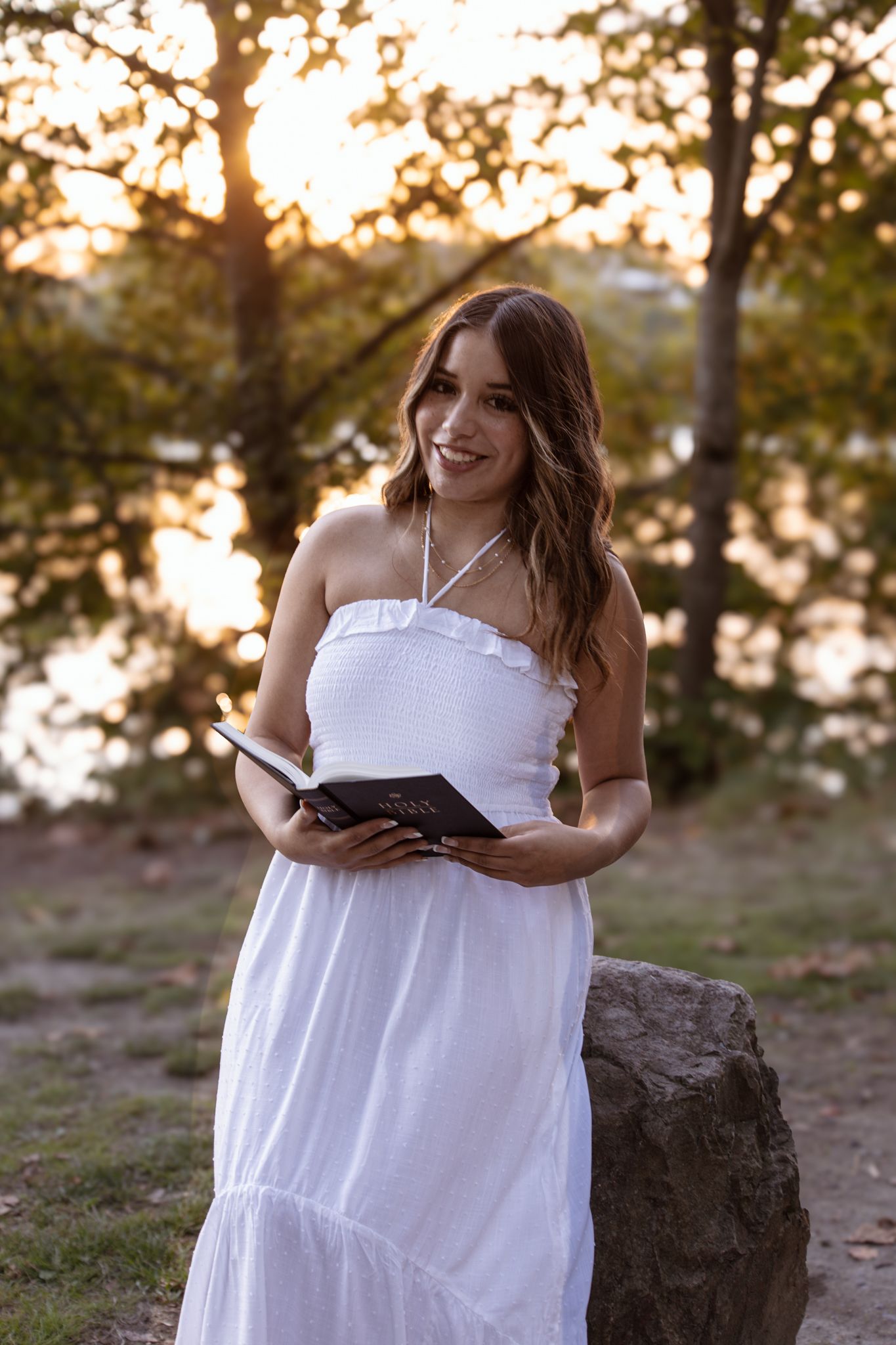 A person in a flowing white dress poses outdoors during golden hour sunset while holding a book.